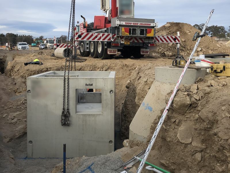 Precast concrete stormwater pit set in an excavation, with a mobile crane behind and a levelling staff in the foreground
